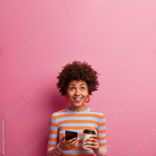 Vertical shot of happy Afro American woman with broad smile, holds disposable cup of coffee and modern smartphone, concentrated above, dressed in casual striped jumper, isolated on pink wall.