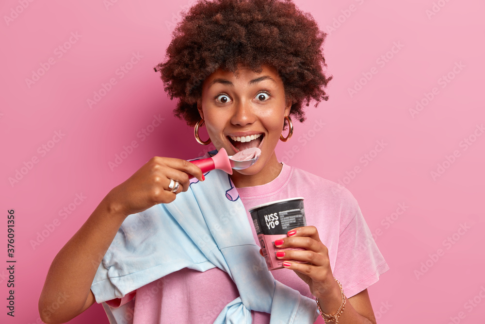 Close up shot of cheerful funny teenage girl with curly hairstyle enjoys eating cold frozen dessert, eats delicious ice cream of strawberry flavor from spoon, stands indoor, pink background. Close up shot of cheerful funny teenage girl with curly hairstyle enjoys eating cold frozen dessert, eats delicious ice cream of strawberry flavor from spoon, stands indoor, pink background.