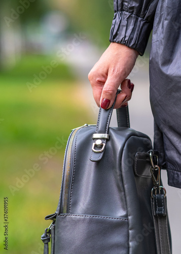 Elegant outfit. Close up of stylish gray backpack in woman s hand. Model holding bag and sitting on the green grass. Female fashion concept.