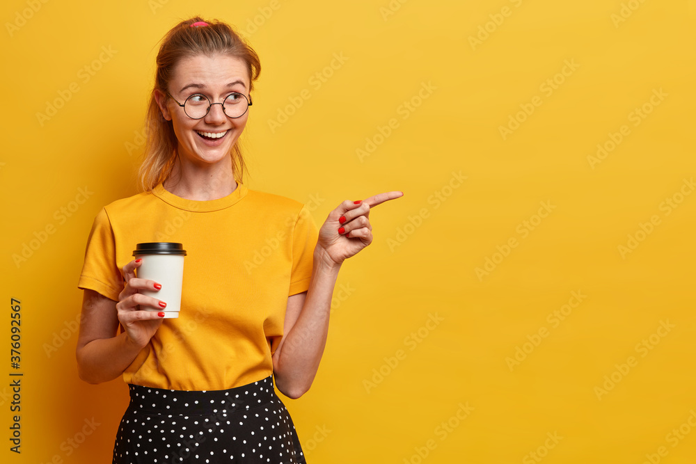 Horizontal shot of good looking millennial girl points right on blank space, invites check out, shows way to advertisement, holds takeaway coffee, wears round big spectacles, yellow t shirt. Horizontal shot of good looking millennial girl points right on blank space, invites check out, shows way to advertisement, holds takeaway coffee, wears round big spectacles, yellow t shirt.