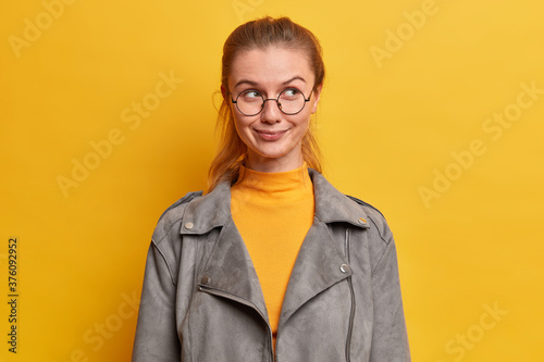 Horizontal shot of pretty young European woman with satisfied expression looks thoughtul aside, has charming smile on face, wears big round spectacles, grey jacket, isolated on yellow background photo