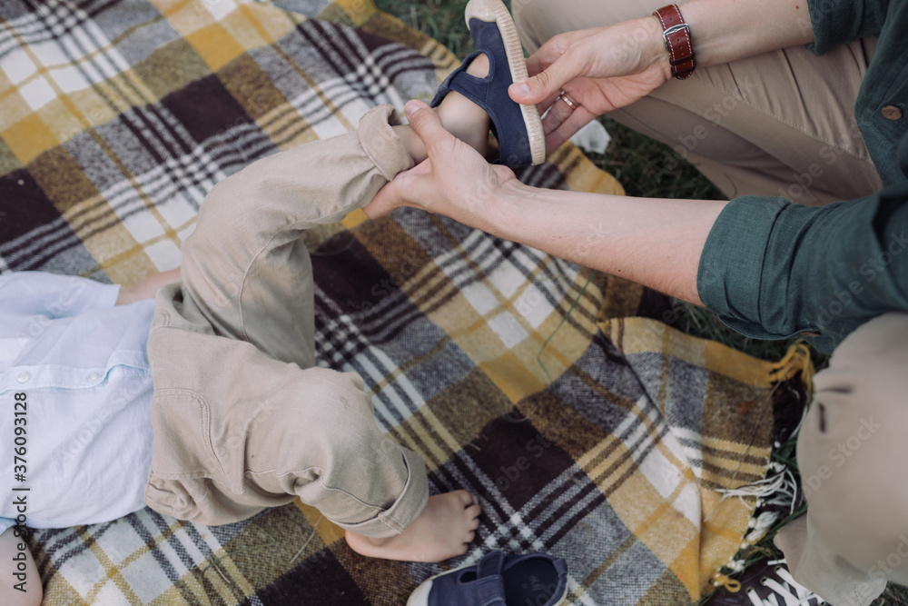 Close up of father putting sandals on to little son on picnic blanket in the park. Family time.