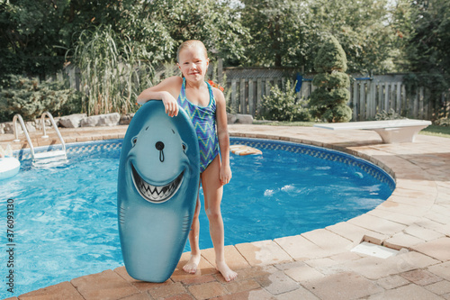 Cute smiling Caucasian girl standing by pool with swim board at home backyard. Kid child enjoying having fun in swimming pool. Summer outdoors water activity for kids.