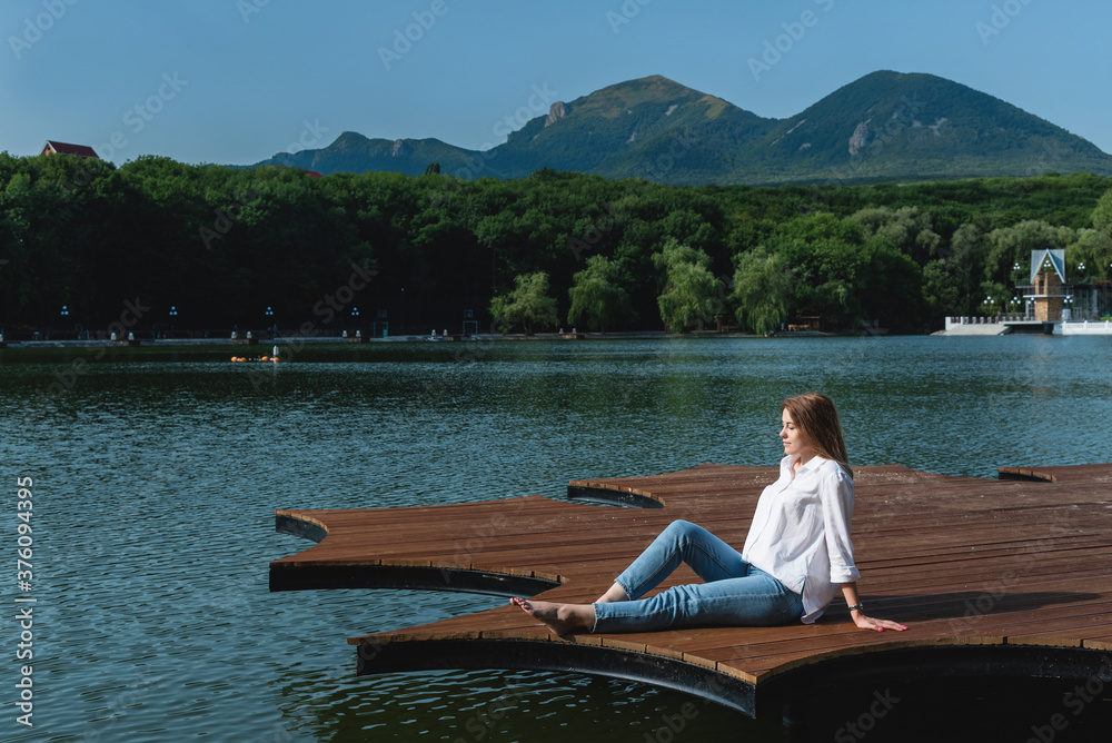 A beautiful and sexy woman, with long hair in a white shirt and jeans, spends time on the shore of the lake, surrounded by forests and mountains. Rest and relaxation concept