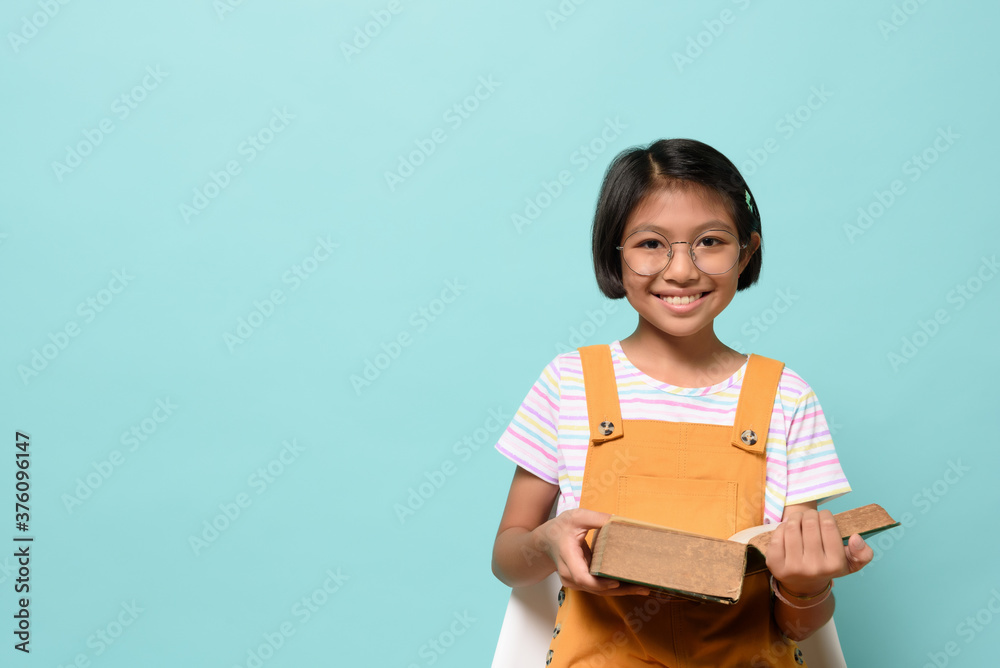 Obraz premium Asian female smile kid wear glasses looking camera and holding dictionary book while sitting chair on pastel blue background in studio. Language Learning of Schoolgirl, learning by heart concept.
