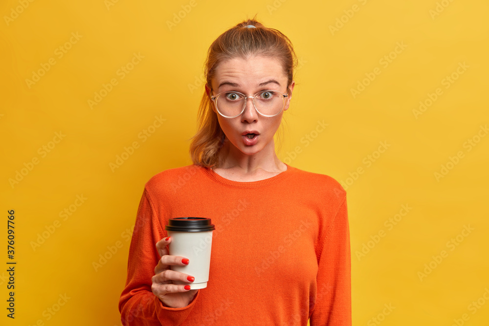 Emotional stupefied female student has coffee break, holds disposable cup of cappuccino, wears big transparent glasses, orange sweater, hears fresh gossip about groupmate, drinks caffeine beverage Emotional stupefied female student has coffee break, holds disposable cup of cappuccino, wears big transparent glasses, orange sweater, hears fresh gossip about groupmate, drinks caffeine beverage