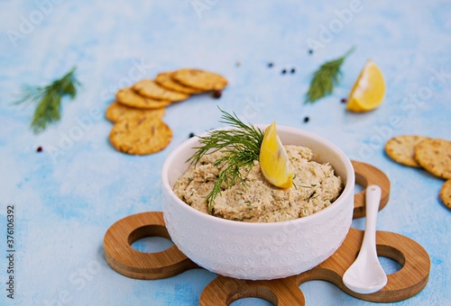 Appetizer, fish pate from mackerel, boiled eggs and onions in a white ceramic bowl on a blue concrete background. Served with savory crackers.