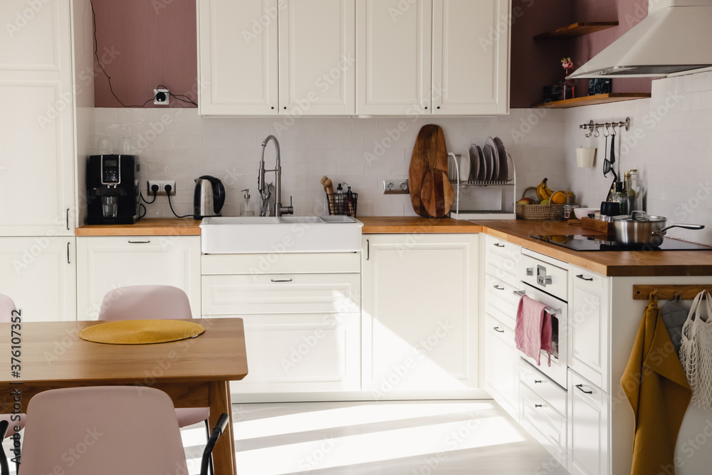 Modern scandinavian style kitchen with white cabinets, wood counter and dining table with sunlight in daytime. Full set of kitchen equipment, pot, electric hob, flipper, fruit. Modern scandinavian style kitchen with white cabinets, wood counter and dining table with sunlight in daytime. Full set of kitchen equipment, pot, electric hob, flipper, fruit.