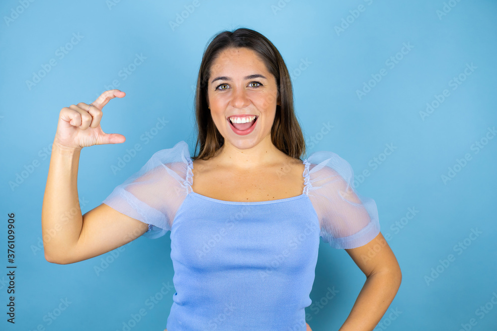Young beautiful woman over isolated blue background smiling and confident gesturing with hand doing small size sign with fingers looking and the camera. Measure concept. Young beautiful woman over isolated blue background smiling and confident gesturing with hand doing small size sign with fingers looking and the camera. Measure concept.