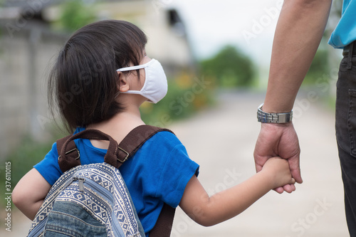 Little girl with backpack wearing cloth face mask hand in her father’s hand waiting for school bus, back to school amid Coronavirus (COVID-19) pandemic. photo