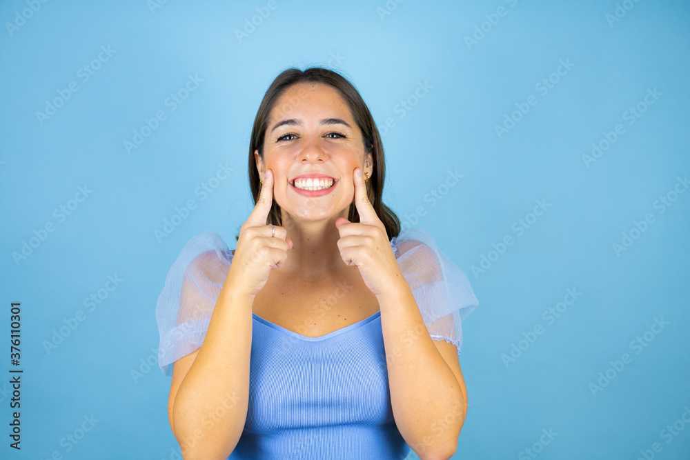 Young beautiful woman over isolated blue background smiling confident showing and pointing with fingers teeth and mouth Young beautiful woman over isolated blue background smiling confident showing and pointing with fingers teeth and mouth