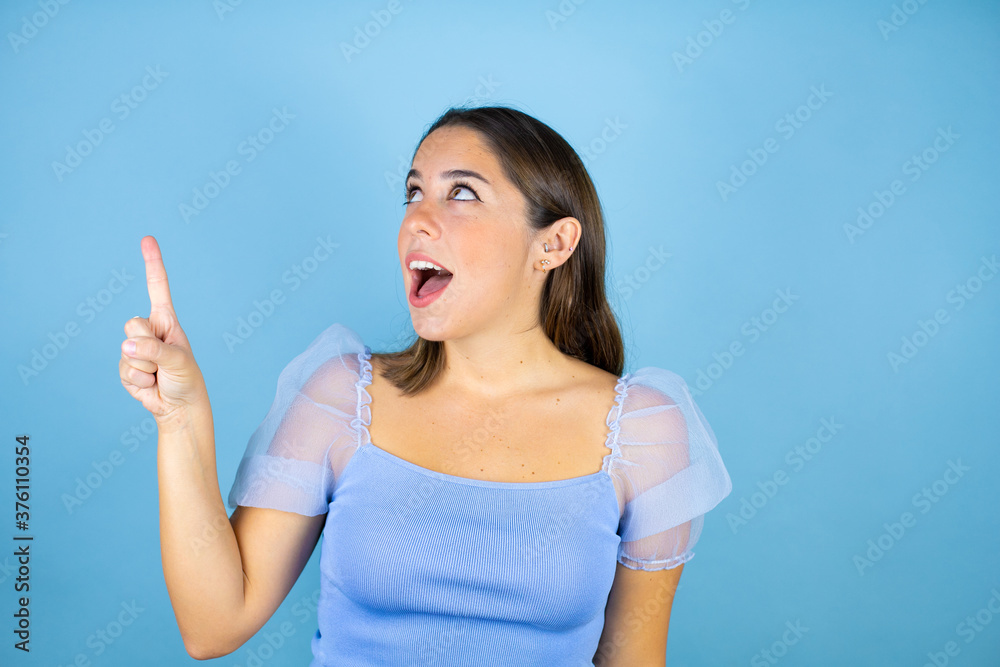 Young beautiful woman over isolated blue background surprised, pointing and looking up. Young beautiful woman over isolated blue background surprised, pointing and looking up.
