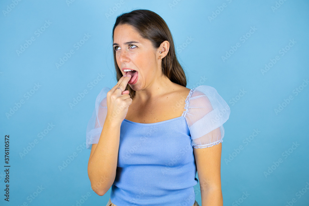 Young beautiful woman over isolated blue background background disgusted with her hand inside her mouth Young beautiful woman over isolated blue background background disgusted with her hand inside her mouth