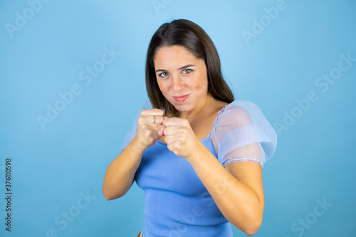 Young beautiful woman over isolated blue background Punching fist to fight, aggressive and angry attack, threat and violence © Irene