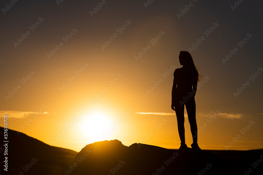 Silueta de mujer joven atractiva con pelo largo al viento al atardecer con el cielo naranja y el sol detrás.