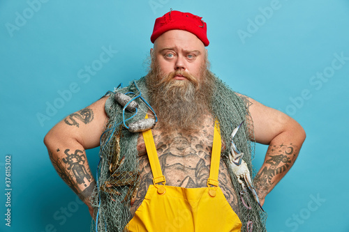 Photo of serious bearded man has confident expression keeps hands on hips, poses with fishing gears, dressed in overalls, red hat, goes agling, poses against blue background. Sea adventure concept photo