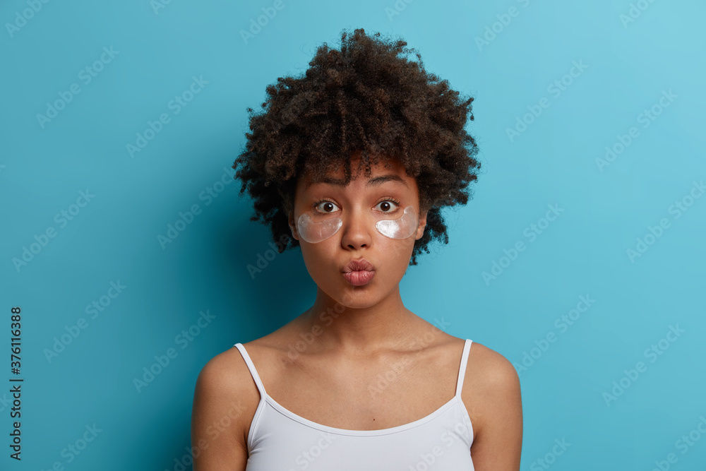Close up shot of young dark skinned woman with Afro hair applies hydrogel silver patches under eyes, reduces puffiness, removes dark circles, cares about complexion, stands indoor. Skin care concept Close up shot of young dark skinned woman with Afro hair applies hydrogel silver patches under eyes, reduces puffiness, removes dark circles, cares about complexion, stands indoor. Skin care concept