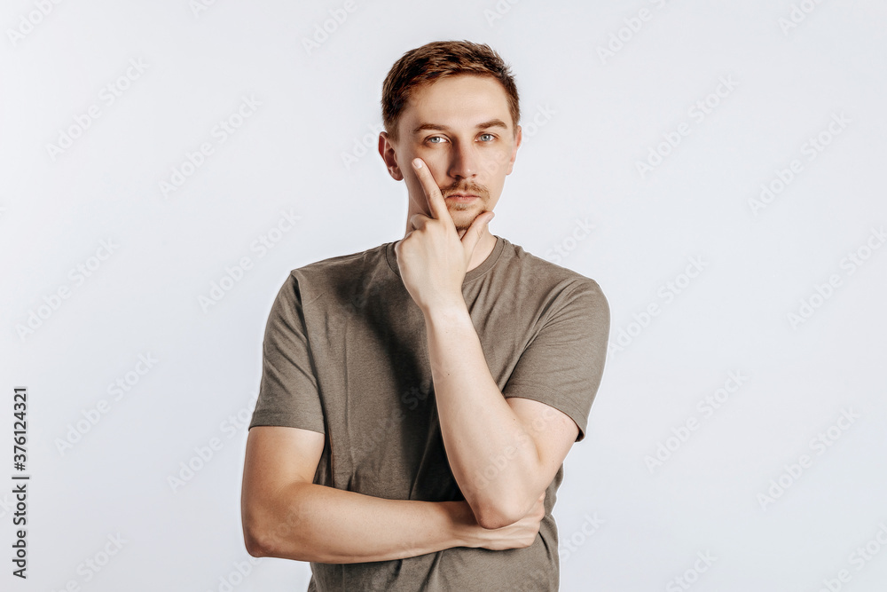 A handsome young man is thinking about a decision and holds his hand to his face. Serious brunette businessman with beard in brown shirt posing over gray background. Place for advertising. A handsome young man is thinking about a decision and holds his hand to his face. Serious brunette businessman with beard in brown shirt posing over gray background. Place for advertising.