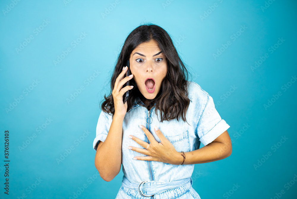 Young beautiful woman wearing a denim jumpsuit over isolated blue background talking on the phone with a worried expression. Young beautiful woman wearing a denim jumpsuit over isolated blue background talking on the phone with a worried expression.