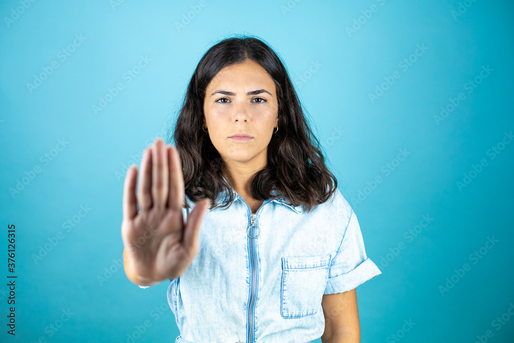 Young beautiful woman wearing a denim jumpsuit over isolated blue background serious and doing stop sing with palm of the hand. Young beautiful woman wearing a denim jumpsuit over isolated blue background serious and doing stop sing with palm of the hand.