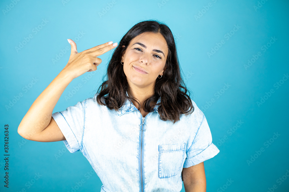Young beautiful woman wearing a denim jumpsuit over isolated blue background Shooting and killing oneself pointing hand and fingers to head like gun, suicide gesture. Young beautiful woman wearing a denim jumpsuit over isolated blue background Shooting and killing oneself pointing hand and fingers to head like gun, suicide gesture.