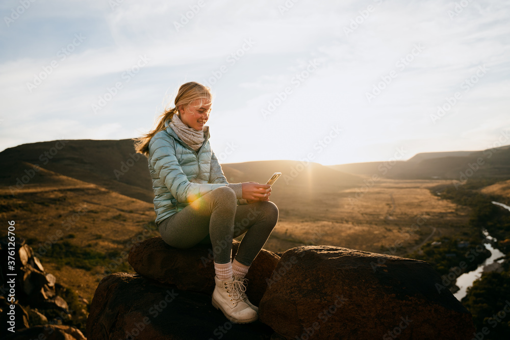 Young caucasian girl scrolling on smartphone while siting on mountain top overlooking river at sunset.