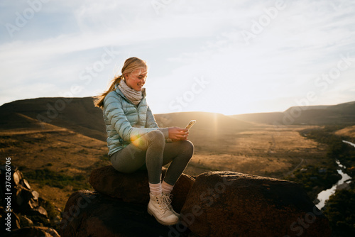 Young caucasian girl scrolling on smartphone while siting on mountain top overlooking river at sunset.