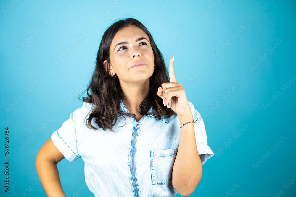 Young beautiful woman wearing a denim jumpsuit over isolated blue background surprised and thinking, having an idea. Young beautiful woman wearing a denim jumpsuit over isolated blue background surprised and thinking, having an idea.