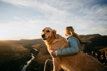 Blonde female sits on top of mountain with arm around golden puppy while looking into the distance at sunset.