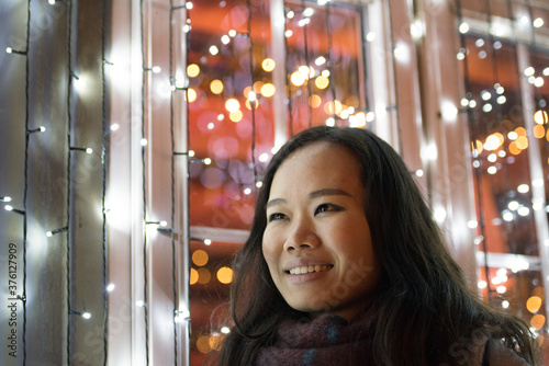 A brunette Asian girl smiles as she looks away in front a magical display of lights on the window of one the cabins in Princes Street Gardens, Edinburgh, UK.