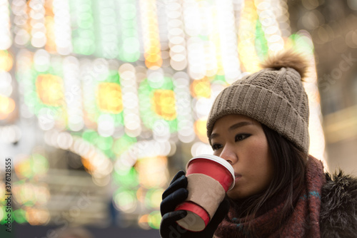 Foto An Asian girl wearing a woolly hat, scarf and gloves drinks from her warm cup of coffee in front of a light display as she looks away in Edinburgh City Center, Scotland, UK