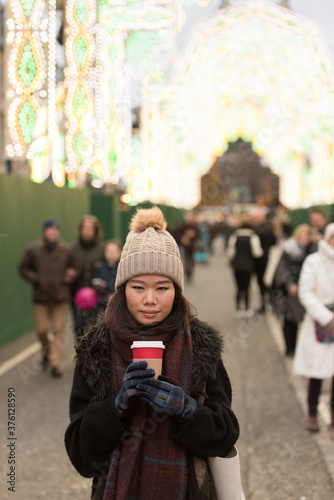 An Asian girl wearing a woolly hat, scarf and gloves holds a warm cup of coffee in front of other pedestrians as she looks at the camera in George Street, Edinburgh City Center, UK.