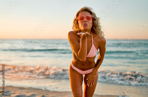 Attractive caucasian woman in a swimsuit and pink glasses blows a kiss while walking at the sunrise by the sea. photo