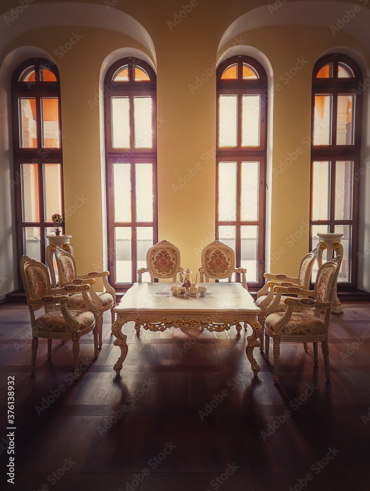 Ancient royalty dining room, medieval furniture style with golden ornate chairs and table near the arched windows. Luxury hosting room background, old wood parquet. Interior architecture details.
