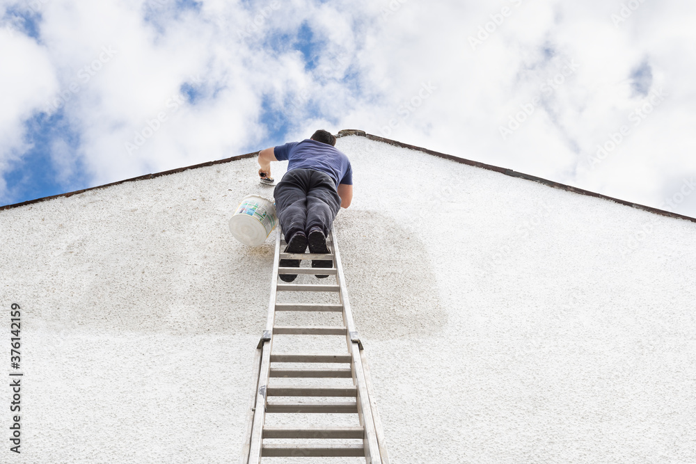 Man stands high up a ladder to paint the side of house a fresh white colour. Man stands high up a ladder to paint the side of house a fresh white colour.