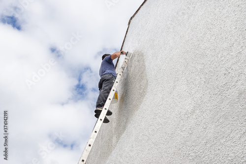 Man up a high ladder painting gable end of pebbledash house.