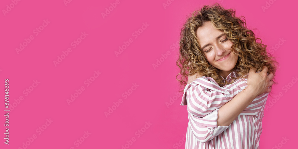 Waiting for a special moment. Portrait of a smiling woman with closed eyes who hugs herself with her arms. Isolated on a pink background.
