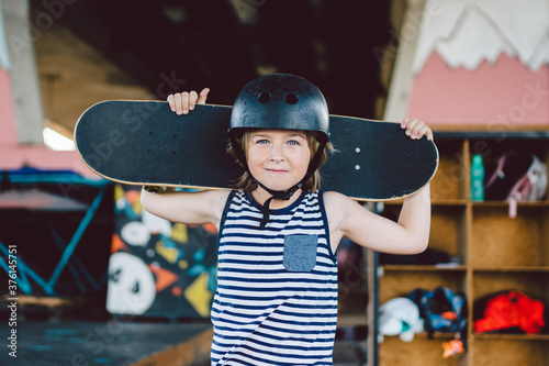 Portrait of handsome caucasian boy athlete skateboarder in protective helmet with skateboard in hands looking at camera on background of skate park. A child and an active hobby, sports and health