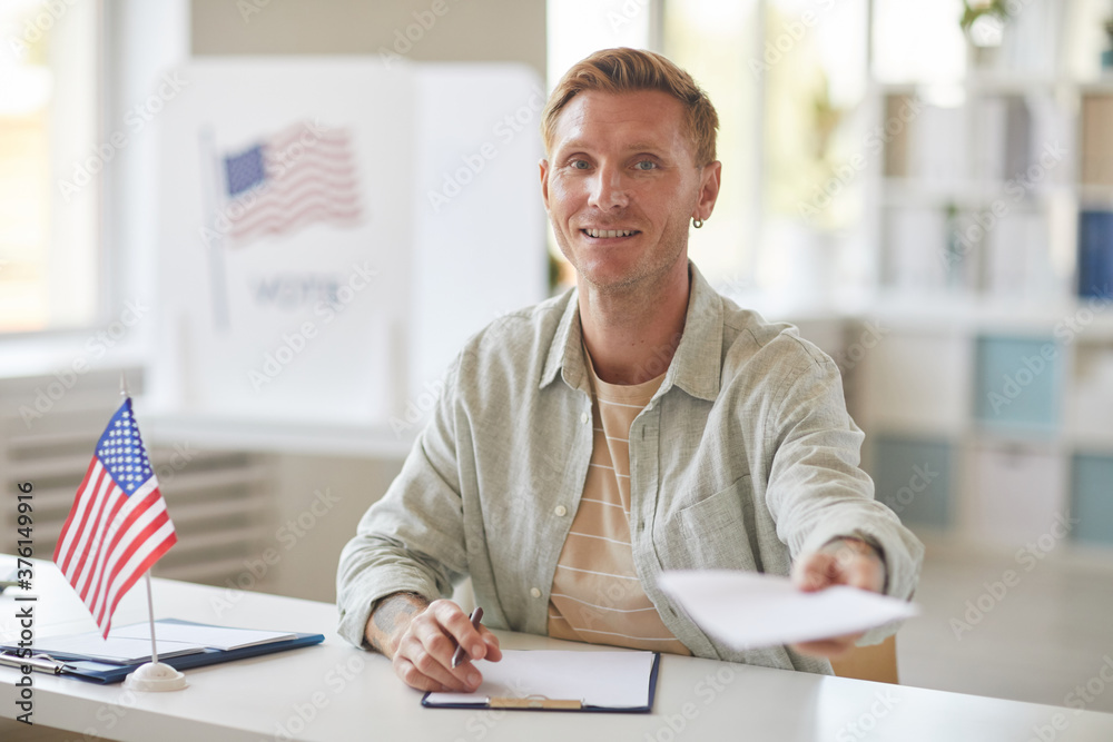 Portrait of smiling modern man handing papers to camera while working at voting station, copy space