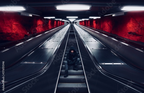 Mysterious man with black hood is sitting on the escalators stair in underground station with rocky structured walls and artificial light sources above, which create beautiful dynamics for the scene. © Zdena Venclik