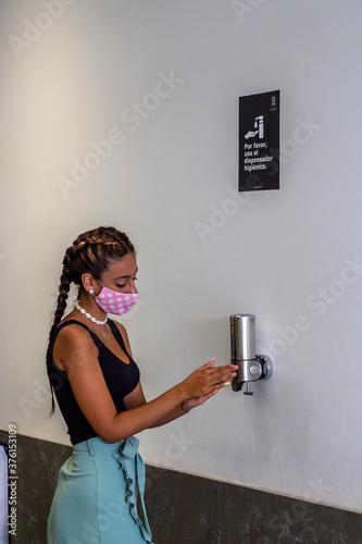 Mujer joven con mascarilla lavándose las manos con gel hidroalcohólico cumpliendo las medidas de seguridad y distanciamiento social de la pandemia COVID-19 en baño publico photo