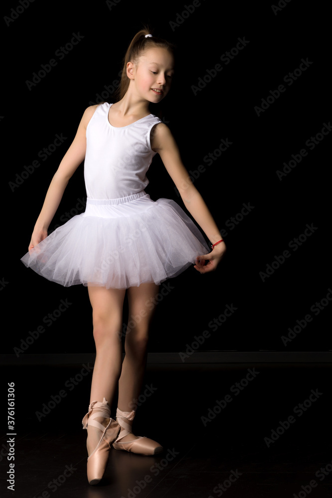 Naklejka premium Cute little girl in a tutu and pointe shoes is dancing in the studio on a black background.