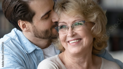 Close up loving young man kissing smiling beautiful mature mother wearing glasses, hugging from back, standing at home, overjoyed happy middle aged woman and adult son enjoying tender moment