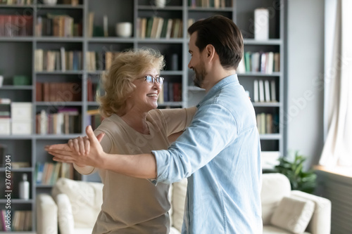 Smiling mature woman wearing glasses dancing with adult son in modern living room at home, young man and happy older mother enjoying leisure time together, moving to music, two generations