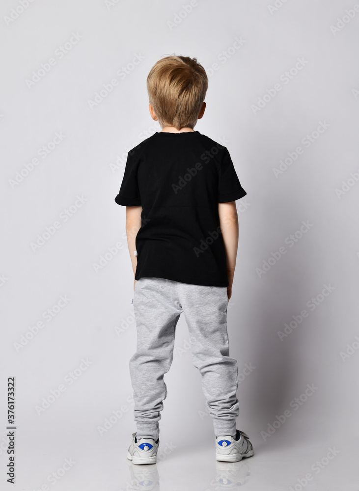 Blond kid boy in black t-shirt, gray pants and sneakers stands back to us with his hands in pockets Blond kid boy in black t-shirt, gray pants and sneakers stands back to us with his hands in pockets