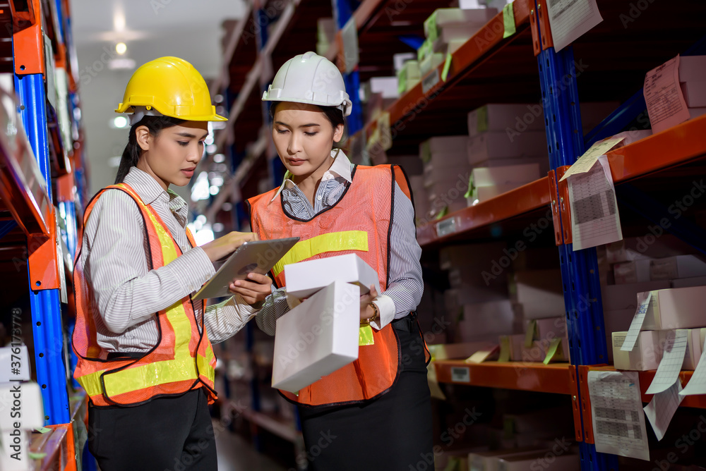 Engineer warehouse woman wearing a hardhat standing cargo at goods warehouse and check for control loading from Cargo freight ship for import and export by report on tablet. Teamwork concept Engineer warehouse woman wearing a hardhat standing cargo at goods warehouse and check for control loading from Cargo freight ship for import and export by report on tablet. Teamwork concept