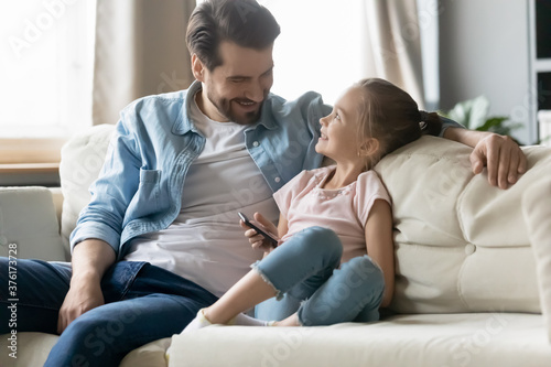 Close up smiling father and little daughter using phone together, sitting on cozy couch at home, happy young dad and adorable child girl enjoying leisure time, weekend in living room