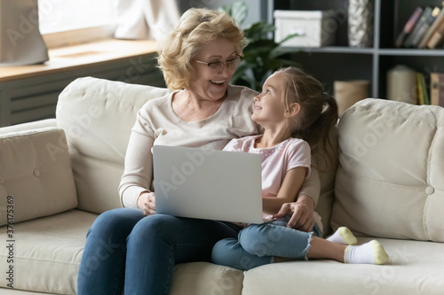 Excited mature grandmother and granddaughter using laptop, having fun together, sitting on cozy couch at home, smiling senior woman wearing glasses and little girl hugging, talking, enjoying weekend