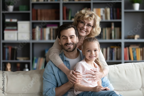Family portrait smiling mature grandmother wearing glasses hugging adult son and little granddaughter sitting on cozy couch in living room, happy mature woman, young man and girl looking at camera