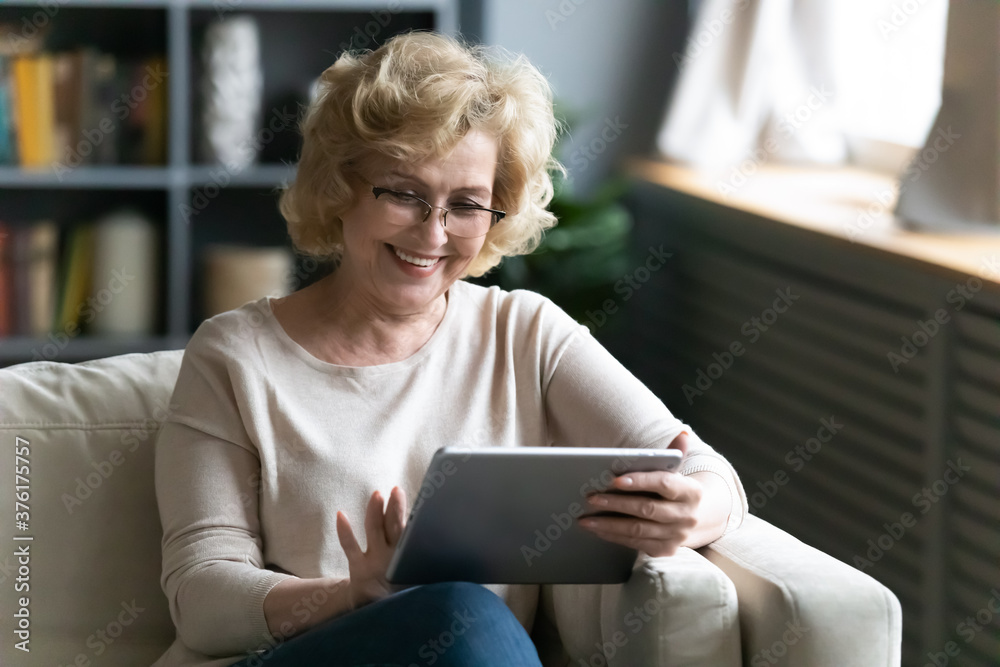Smiling mature woman wearing glasses using computer tablet at home, positive overjoyed middle aged female browsing apps, shopping or chatting online, sitting relaxing on cozy couch at home Smiling mature woman wearing glasses using computer tablet at home, positive overjoyed middle aged female browsing apps, shopping or chatting online, sitting relaxing on cozy couch at home